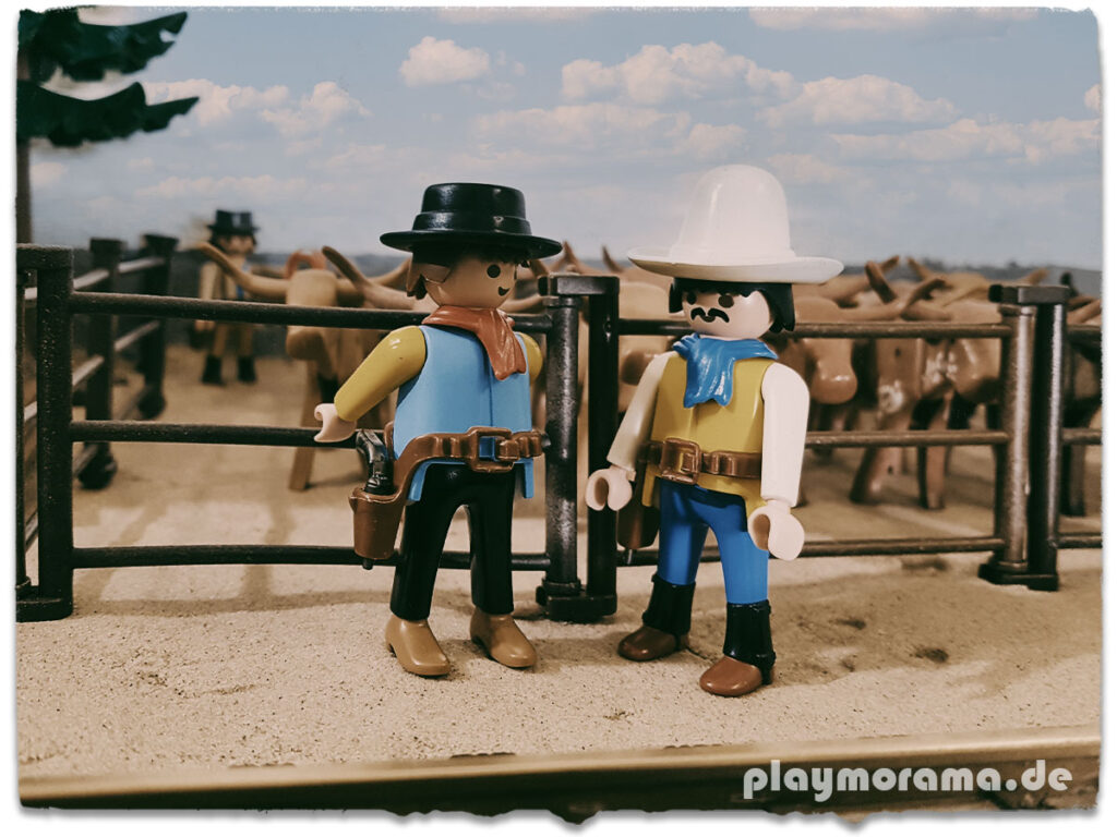 Vor der Western-Station sind Longhorn-Rinder in den Stallungen untergebracht, während zwei Playmobil Cowboys sich um sie kümmern und sich auf den bevorstehenden Viehtrieb vorbereiten.