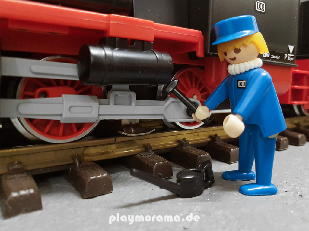 Der Dampflokmechaniker mit gelben Haaren und Halskrause hält einen Hammer in der Hand und arbeitet an der Lok.