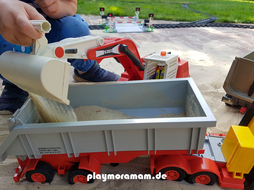 Playmobil Baustellenlaster wird von einem Bagger mit Sand beladen. Ein Junge spielt mit Playmobil draußen auf der Terasse.