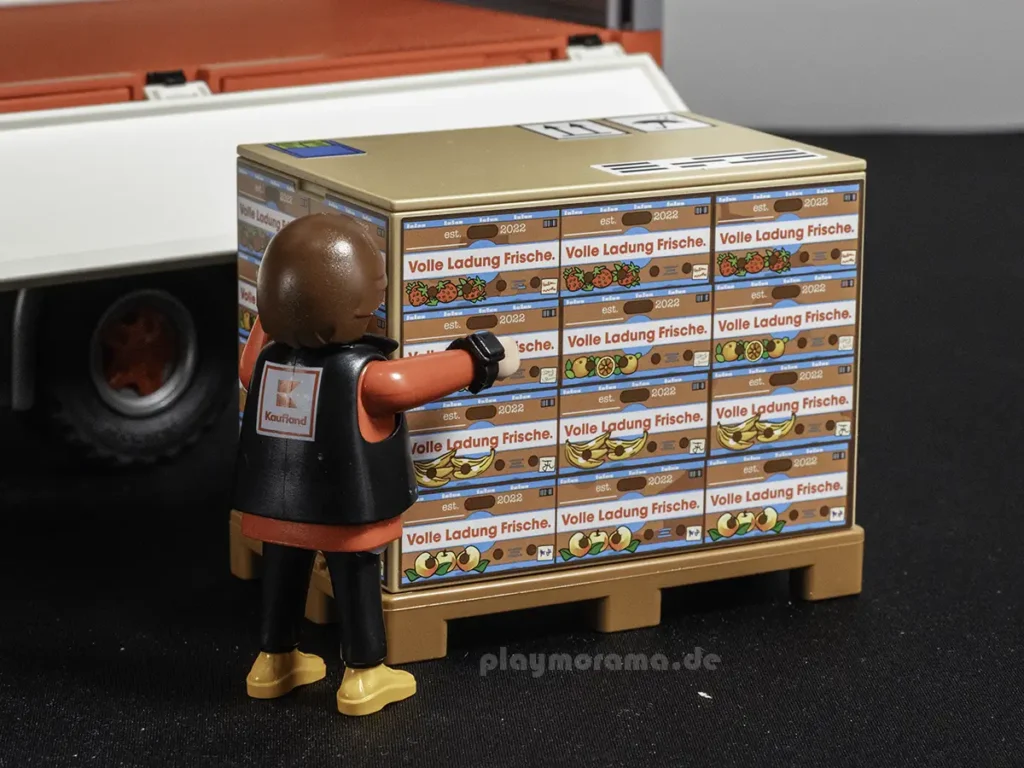 Die Playmobil-Figur trägt den Aufdruck „Kaufland“ auf der Weste. Die Box auf der Palette ist mit „Volle Ladung Frische“ beschriftet. Die Sticker zeigen detailreiche Obstkisten.