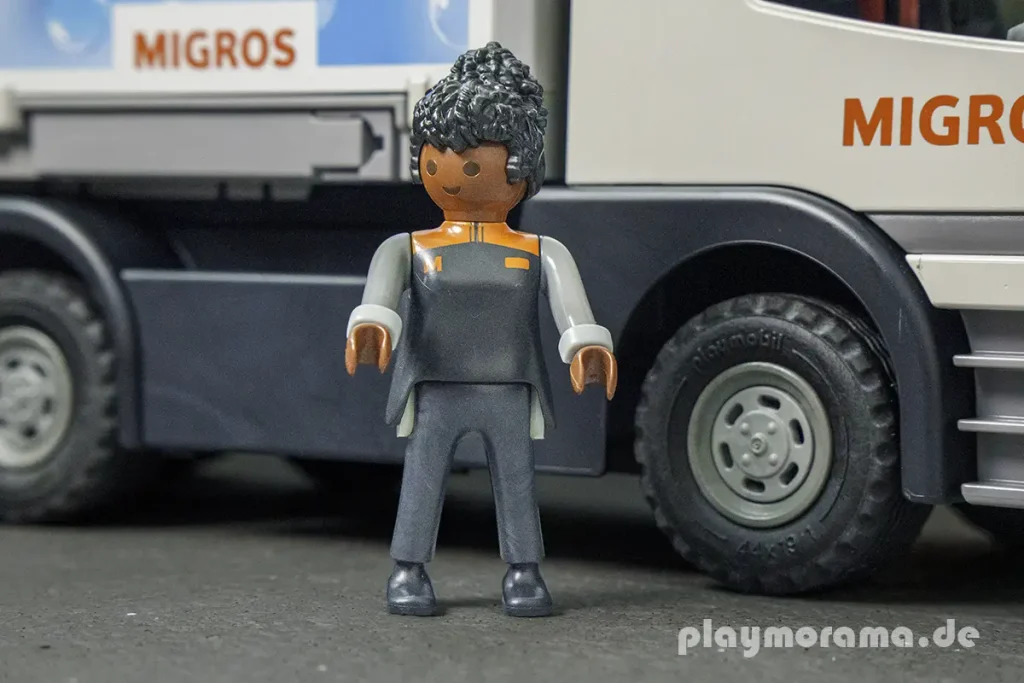 PLAYMOBIL-Migros-Fahrerfigur in grauer Arbeitskleidung mit orangefarbenem Kragen vor dem LKW